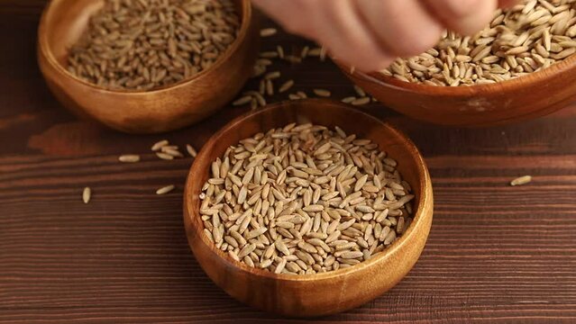 hand pours rye grains into a wooden bowl on a brown wooden background, side view, close up