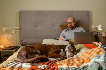 sick man in bed working, accompanied by dog sleeping on feet