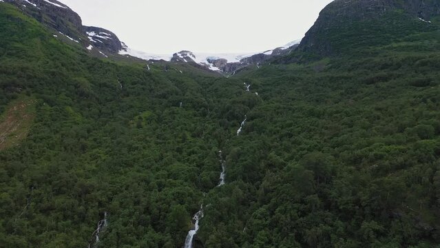 Several river streams flowing from the melting Myklebust glacier through lush dark green forest in Olden Norway - Aerial flying up hillside while looking at river stream with glacier in top of frame