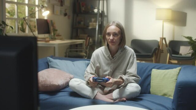 Medium shot of young woman sitting with legs crossed on sofa at home and smiling while playing video game on TV