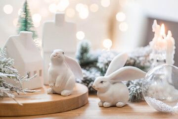 Fototapeta na wymiar Beautiful holiday card. The concept of a happy new year and Christmas 2023. Christmas trees, a white rabbit and Scandinavian white houses on a wooden table in a cozy living room.