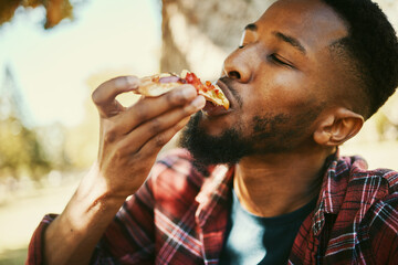 Fast food, hungry and black man eating pizza for delicious and yummy lunch break in park. Gen z, food and hunger of young person enjoying carbs pizza slice meal with satisfied face.