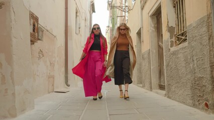 Young blonde and brunette haired women in colorful outfits with fashionable handbags stroll along narrow Venetian street between historical buildings