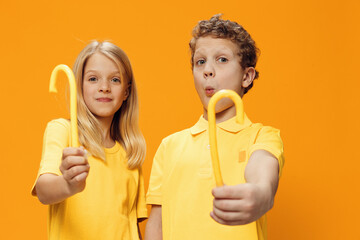  happy children brother and sister, stand in bright yellow clothes with yellow lollipops in their hands and the girl makes a funny emotional face. Studio horizontal photography on orange background