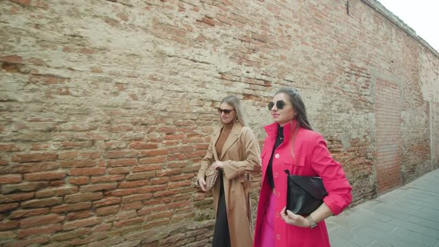 Young girl friends with long brunette and blonde hair holding purses and wearing accessories talk walking along shabby brick wall in Venice