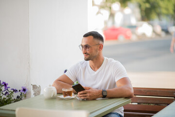 a happy man in a white T-shirt, talking on a cell phone, sitting at a table in a cafe on the street.A man has breakfast and drinks tea in a cafe, Freelance office business