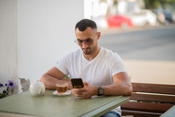 a happy man in a white T-shirt, talking on a cell phone, sitting at a table in a cafe on the street.A man has breakfast and drinks tea in a cafe, Freelance office business