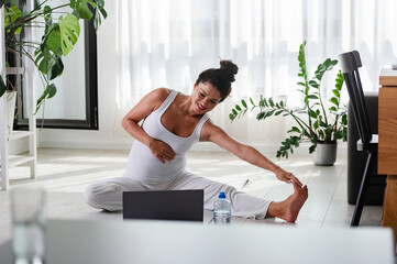 Pregnant woman exercising at home with laptop. Expectant mother doing prenatal video training class indoors. Female exercise during pregnancy. Online fitness class on digital devices 