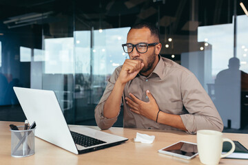 Businessman coughing at work in office, african american man in glasses and shirt sick at workplace using laptop at work.