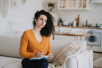 Pensive young adult arabic woman in orange blouse sitting on cozy sofa holds notebook looks aside...