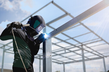 Male worker using steel pole welding machine, blue sparks from welding, wearing mask and gloves for...