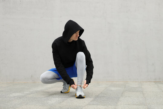Runner tying shoelaces against a gray concrete wall. Athlete in warm sportswear.