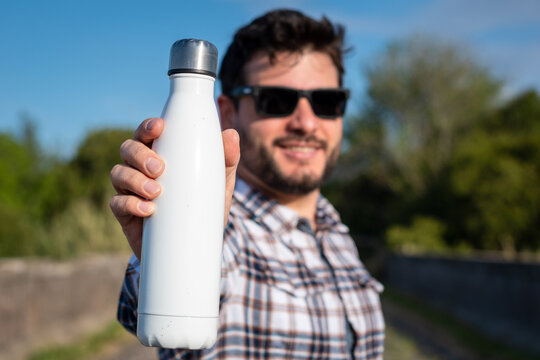 Hombre apuesto y sonriente mostrando botella met&aacute;lica en blanco, para anuncios o propagandas. Fotograf&iacute;a con enfoque selectivo en la botella