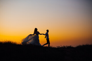 Fototapeta na wymiar silhouette of a cheerful couple, the bride and groom in a wedding dress, laughing and holding hands, run across the field.