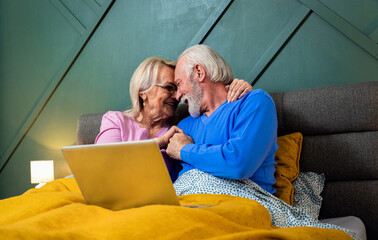 Smiling senior couple lying in bed in the bedroom using laptop. - Powered by Adobe