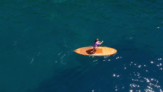 Young woman on a stand up paddle board SUP rawing among beatyful rocks. Clear blue sea water surrounds her. Aerial slowmotion video