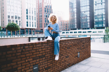 Cheerful ethnic woman using smartphone sitting in street during coffee break