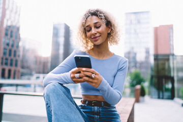 Fototapeta premium Smiling ethnic woman using smartphone sitting on wall on street