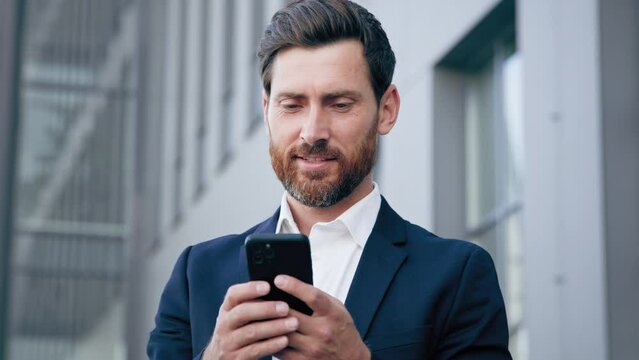 Young successful businessman holding smartphone standing in city business center uses phone writes text message smiling man manager using mobile application for telephone remote communication concept