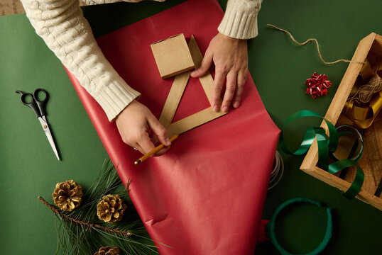 Top view woman's hands using a wooden triangular ruler to draw on red gift paper, calculating the amount of packaging materials needed to wrap Christmas present, New Year or any other holiday event