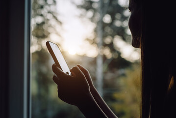Close-up of young caucasian woman sits with phone sitting on cozy window sill and enjoys scenic view on beautiful autumn forest during sunset or sunrise. Recreation and escaping to nature
