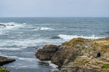 Fototapeta na wymiar Middle-aged couple of man and woman contemplating the Atlantic Ocean sitting on a ledge of rocks with moss and lichen watching as the waves of the sea go to the coast under a gray sky