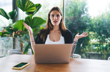 Young female student with laptop shrugging in cafe