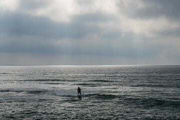 Obraz premium Young surfer man standing on his board submerged in the sea water under a cloudy sky about to rain and the water behind him illuminated with rays of the sunset sun coming out from between the clouds
