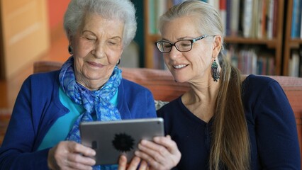 Happy elderly senior woman and mature woman look at tablet computer while drinking tea on a couch in a living room.