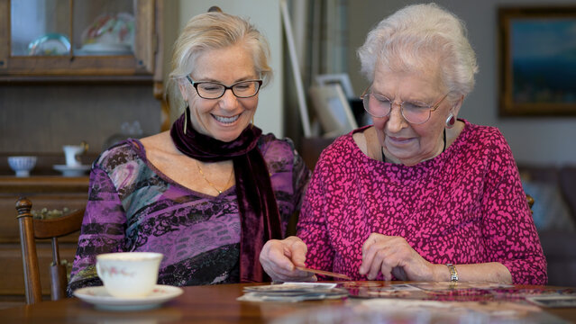 Senior elderly smiling happy woman looking at old photos and remembering memories with daughter at the dining room table.