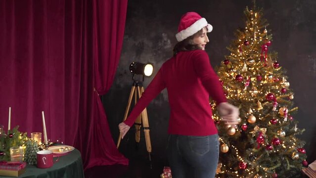 Woman in red sweater and Santa's hat dancing near the Christmas tree