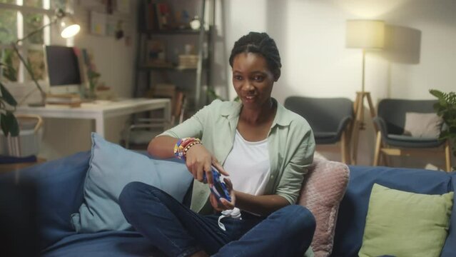 Medium shot of African American woman sitting on couch at home and playing exciting console game