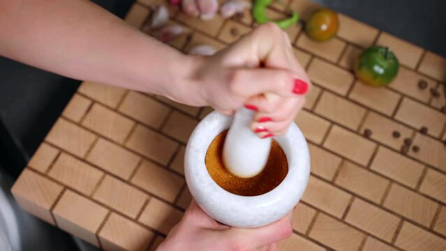 Preparation of aromatic spices mix for meat, vegetables or fish seasoning, person hand using pestle for crushing assortment of spices in stone mortar bowl on professional wooden board. Preparing