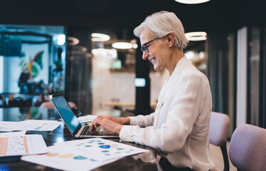 Fototapeta premium Cheerful aged businesswoman writing email on netbook while working
