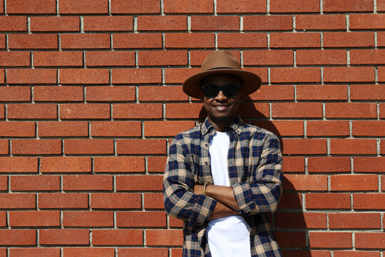 Young black man wearing wide brimmed fedora hat, sunglasses and checked shirt, posing over red brick wall background with copy space. Tanzania born hipster guy leaning on the wall.