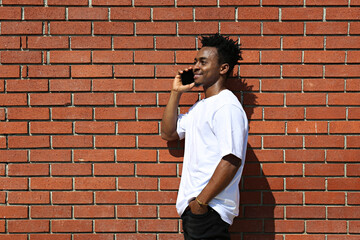 Fototapeta na wymiar Young black man on the street seating on the curb with his phone and texting. Portrait of a guy wearing a casual outfit leaning on the red brick wall with copy space for text. Background.