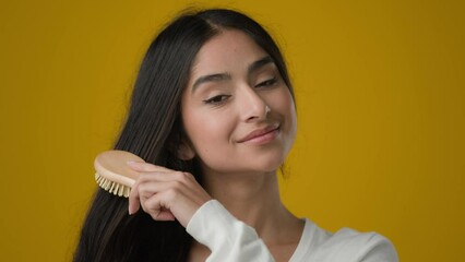 Indian 20s woman ethnic girl lady female brushing combing hair looking at camera doing hairstyle with comb hairbrush get ready everyday routine shampoo conditioner result in yellow studio background