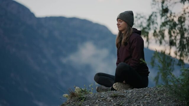 Young woman in hiking clothes meditating on a rock with high mountains in the background