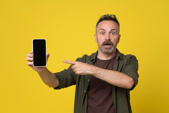Surprised mature man pointing index finger to his smartphone with black screen on yellow background. Handsome man dressed in green shirt holding and showing screen of smartphone in hand - Powered by Adobe
