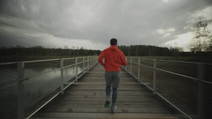 Male in orange hoodie running through national park across wooden bridge in wilderness in the cold fall weather. Sportsman does a fitness workout jogging on a timber footbridge in the countryside. 