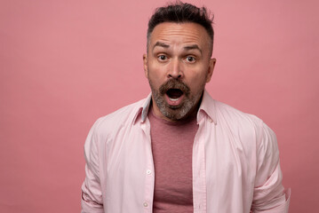 Astonished with open mouth and big surprised eyes attractive mature bristle man isolated on pink pastel background dressed in a light pink shirt