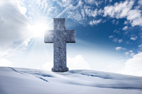 Large stone Christian cross in a mountain landscape with snow against a clear blue sky with clouds, sun rays and copy space.