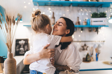 Cheerful woman cuddling little child in kitchen while cooking