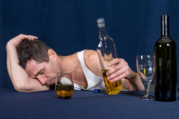 Lonely drunk man in homemade white T-shirt lies with his chest on table and sleeps. He holds bottle of alcohol in his hand. Alcoholism as disease.