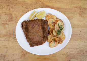 Top view of traditional milanesa, breaded fried steak, with chips and lemon, in a white dish on the wooden table. 