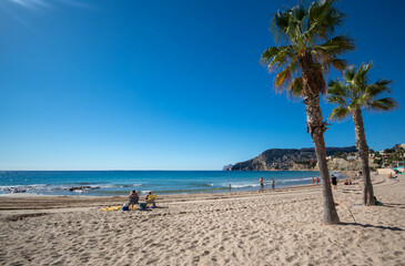Fototapeta na wymiar Personas relajándose en la playa del pueblo de Calpe con un par de palmeras plantadas en la arena bajo un precioso cielo azul soleado de otoño.