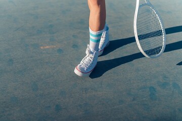 Female legs in blue athletic socks and sneakers with a tennis racket on a court