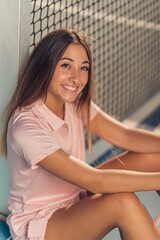 Vertical shot of a young female in pink athleisure posing sitting on a tennis court