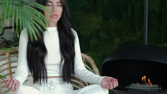 Beautiful woman sits cross-legged on chair in room with greenery and fireplace, practices meditation with her eyes closed, feels inner harmony and balance, improves self-awareness, no stress concept