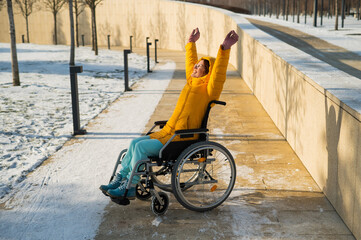 Happy woman in wheelchair happily pulls her hands up while walking in winter park. 
